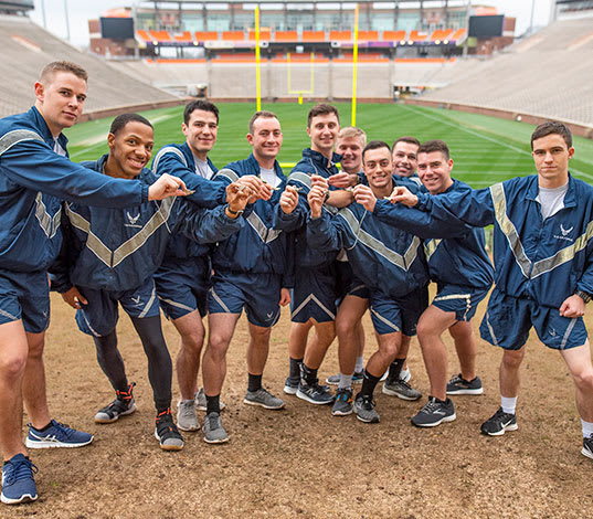 Male air force ROTC cadets in physical training attire hold out their wing pins and smile in Memorial Stadium.