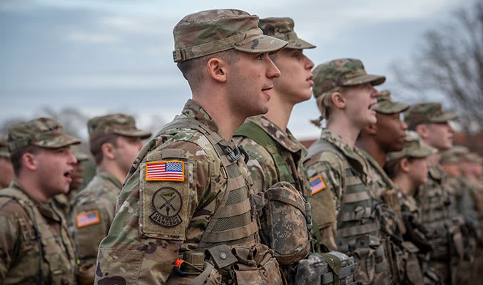 Current Clemson ROTC cadets wearing camouflage uniforms stand in line on Bowman Field speaking in unison.