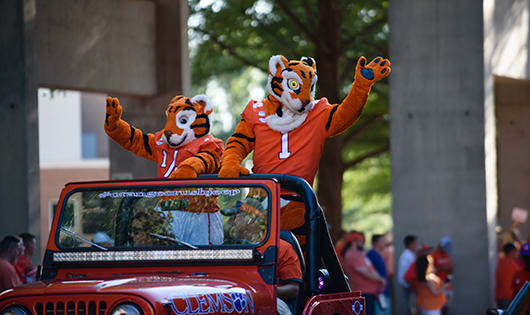 Two Tiger mascots wave to fans and ride in an orange jeep in the shade of Memorial Stadium during the First Friday parade.