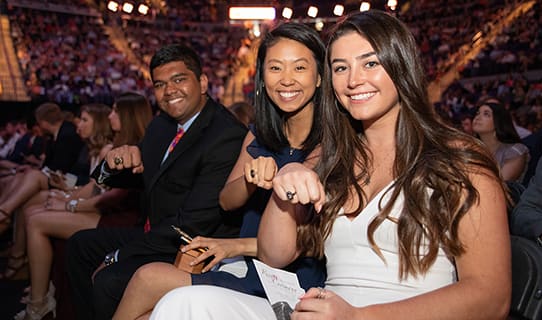 Three seniors in a crowded row of students smile and show off their new Clemson rings during the ring ceremony.