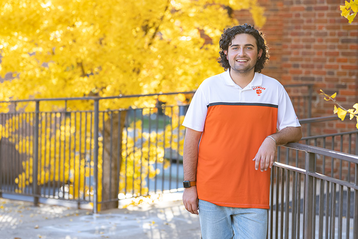 A male student wearing light-wash blue jeans and an orange and white Clemson polo leans against a metal railing outside a brick academic building.