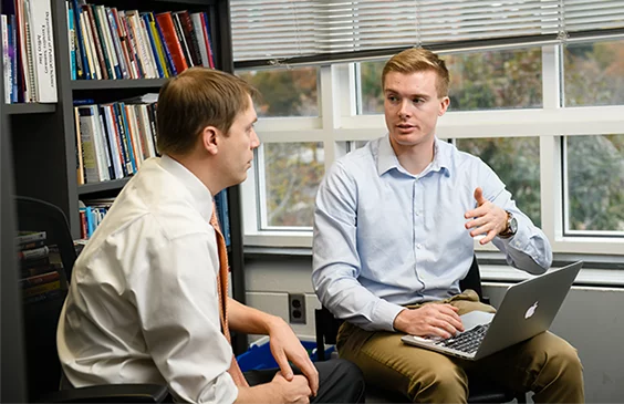 A male student wearing a light blue button-down shirt and khakis sits in a chair with his laptop and talks to a male professor, who is also seated.