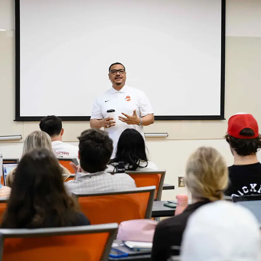 A man wearing a white polo and holding a coffee cup stands in the front of a classroom and speaks with a group of students who sit in orange chairs at classroom tables.