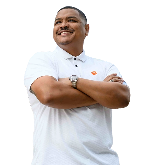 A smiling man in a white polo with an orange Tiger Paw logo stands with his arms crossed.