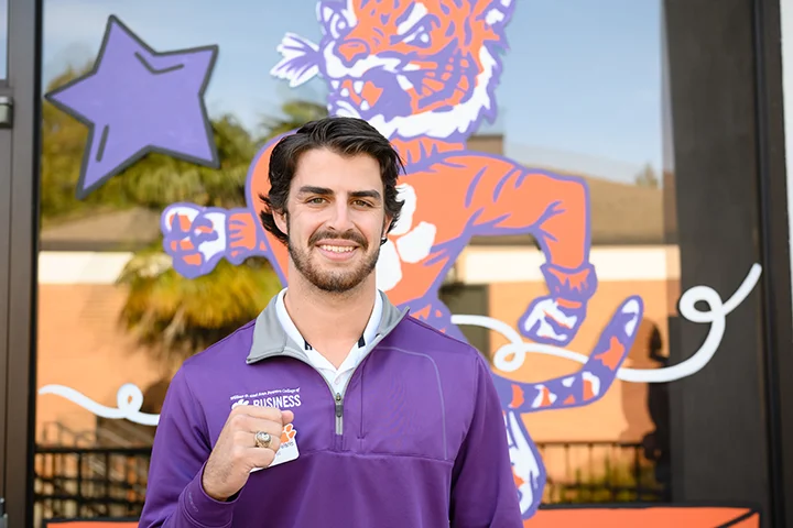 A male student in a purple jacket stands in front of a window painted over with the Clemson Tiger and a purple star.