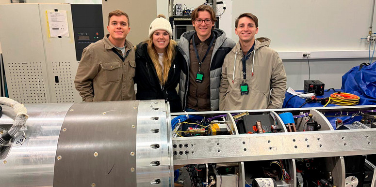 Three male students and a female student stand in an industrial research laboratory behind a large cylindrical scientific instrument with exposed wiring and mechanical components.