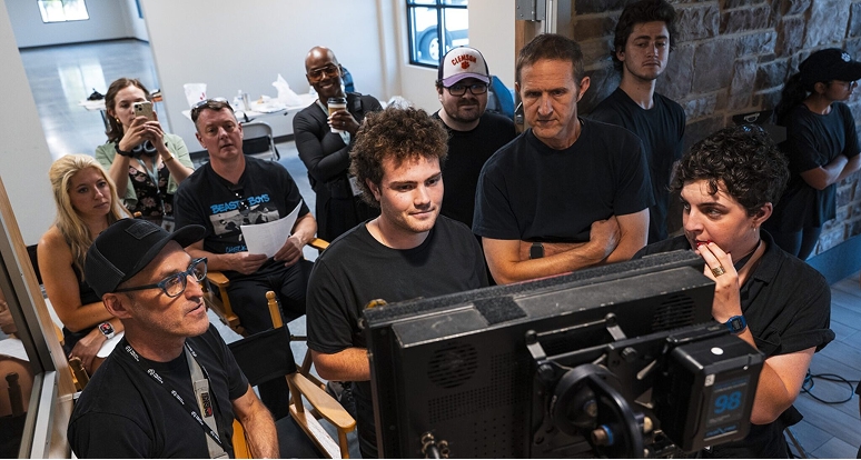 A group of film crew members, all wearing black, gather closely around a large rectangular monitor on a set. Several people sit in director’s chairs while others stand behind them, all focused on the screen. 