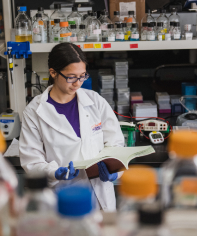 A female science student wearing glasses reads a book in her lab with glass bottles lining the shelf behind her.