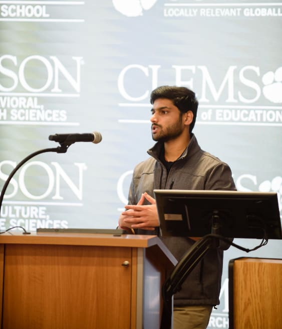 Male student speaks, standing behind podium with College of Education logo on screen behind.