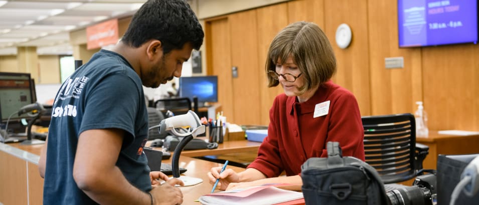 Middle-aged female librarian at the Cooper Library help desk assists male college student with locating a book.
