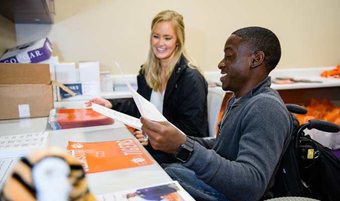 A male student in a wheelchair and a female adviser look though paperwork with a Clemson folder on the desk.