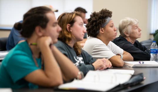 Four diverse students sit at a table, listening to a presenter with books and notes in front of them.