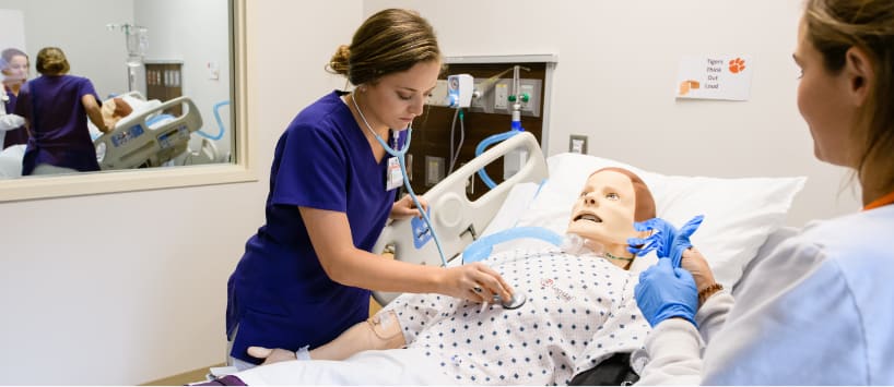 Female nursing student listens to the heartbeat of a mock patient in a lab setting. 