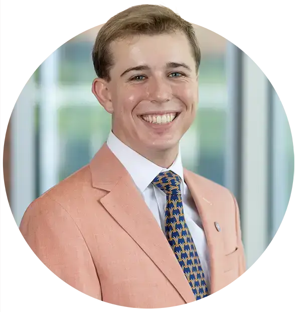 A headshot portrait of a male student wearing a white button-down shirt, a tie with a blue and yellow pattern, and a peach blazer.