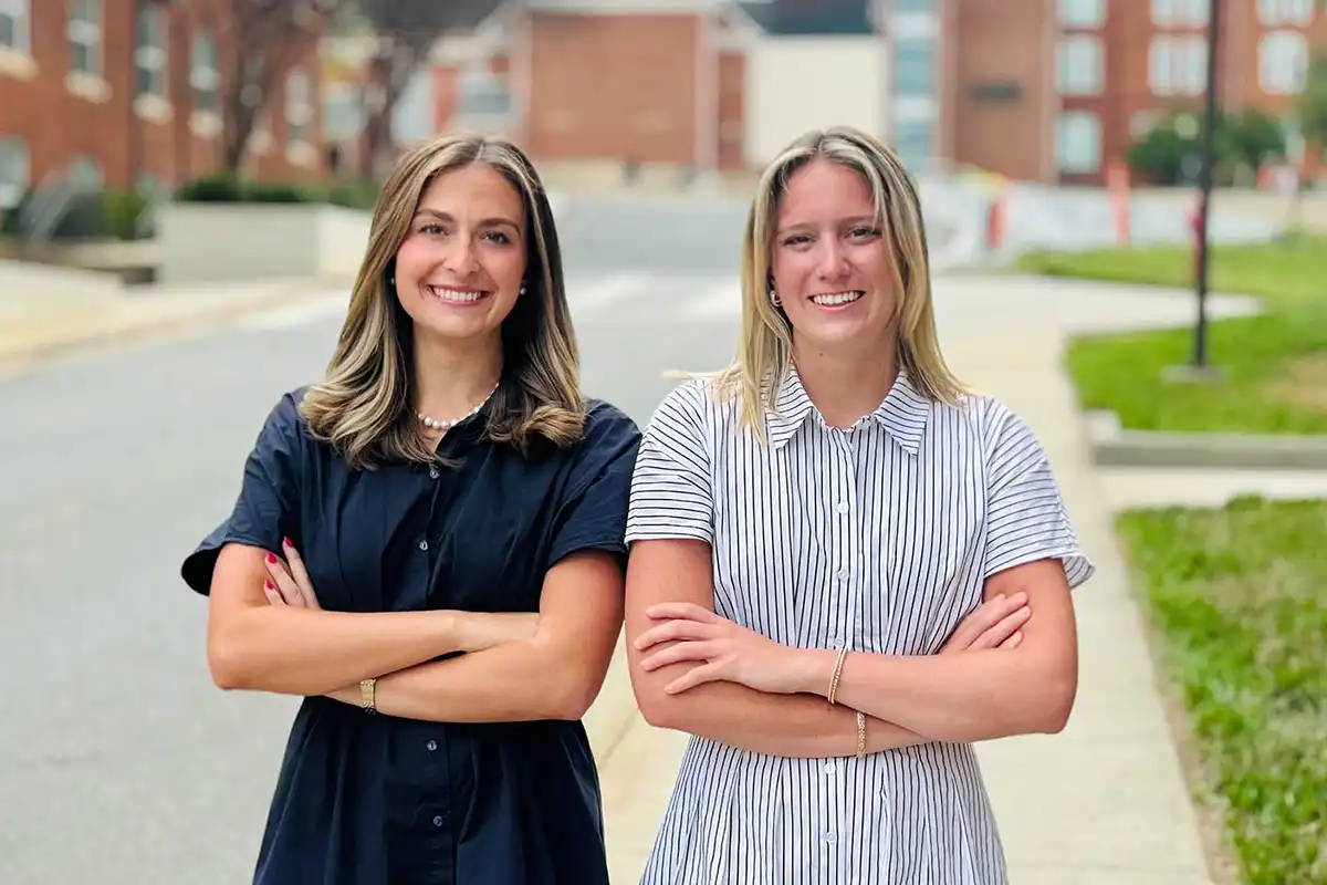 A female student wearing a navy blue dress and a female student wearing a white dress with vertical blue stripes stand outdoors and smile next to each other with their arms folded with brick campus buildings in the background.