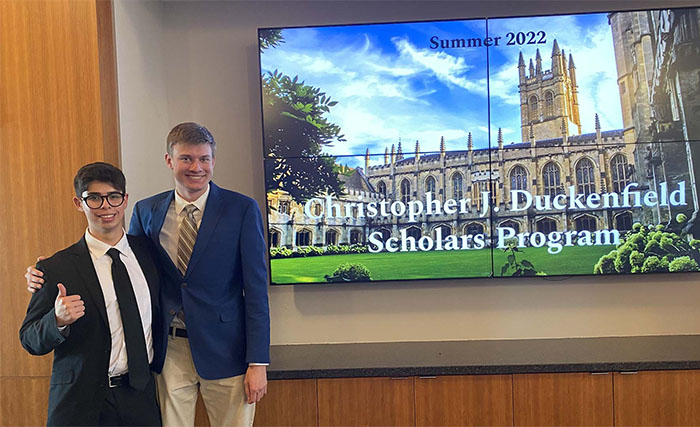 Two male students in formal business attire pose in front of a screen that displays a Gothic architectural academic building and reads, Summer 2022: Christopher J. Duckenfield Scholars Program.