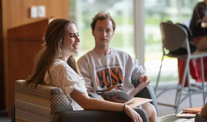 A female student sits in a chair in a student lounge with a male student.