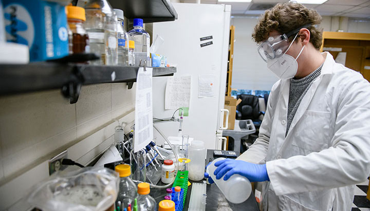 Student Luke Broughton, wearing a white laboratory coat, blue gloves, protective goggles and a white N95 face mask, pours a solution in a laboratory. 
