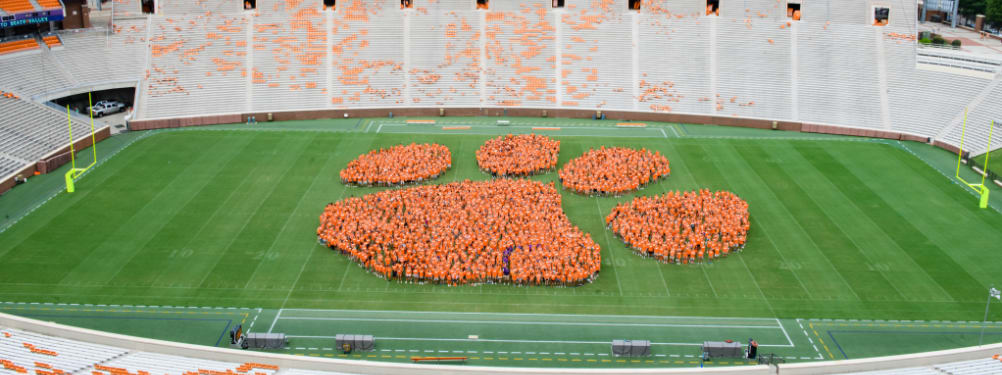 Clemson students in orange t-shirts stand in the shape of a tiger paw on the football field in Death Valley.