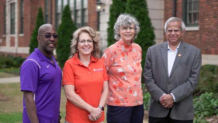 A man in a purple polo, a woman in an orange polo, a woman in a floral orange top and a man in a gray suit jacket stand together in front of a brick campus building. 