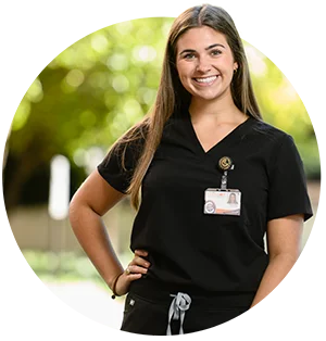 A smiling female student in black medical scrubs with a Clemson ID tag hanging from her collar stands in front of distant trees.