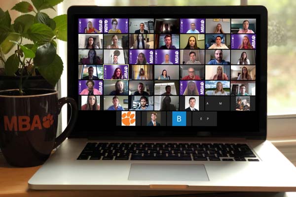 A Mac computer screen shows a Zoom meeting with dozens of participants during Clemson’s MBA program Orientation. Next to the laptop sits a black mug that reads “MBA” in orange type with an orange Clemson Paw.