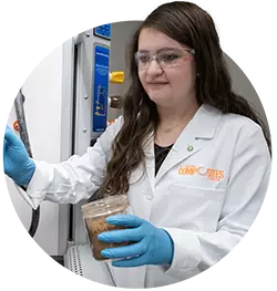 A female student wearing a white lab coat and blue gloves holds a jar in a laboratory setting. 