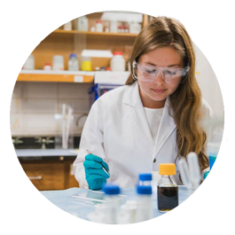Graduate student Riley Rapert, wearing a white lab coat and protective goggles, takes notes in a laboratory.