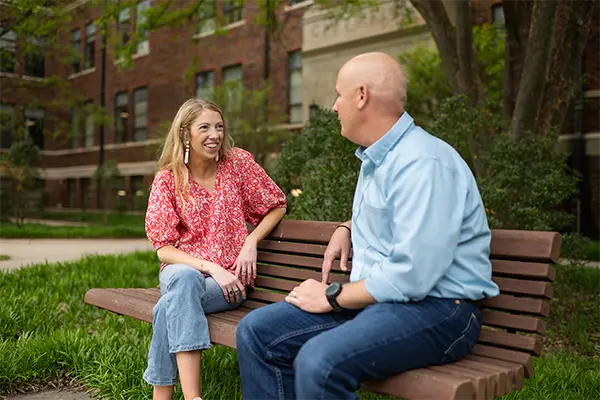 A female counselor wearing jeans and a pink floral top sits on a wooden bench with a male professor wearing jeans and a light blue button-down top. 
