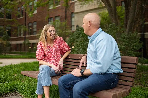 A female counselor wearing jeans and a pink floral top sits on a wooden bench with a male professor wearing jeans and a light blue button-down top.