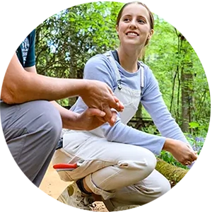A female student wearing a light purple long-sleeved T-shirt and cream overalls kneels on a dirt walking path in a garden and speaks to another woman wearing gray pants, a teal polo and a. straw hat.