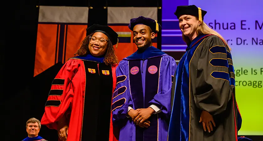 A male doctoral graduate wearing purple academic regalia poses with two female professors — one in red regalia and the other in gray regalia — during the doctoral hooding ceremony.