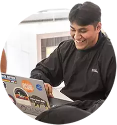 A male student wearing a black sweatshirt looks at his laptop computer that is covered with stickers.