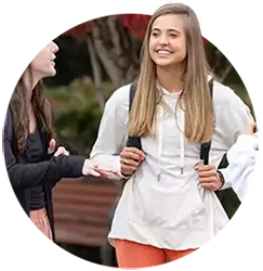 A female student wearing a white sweatshirt and orange pants speaks with another female student.