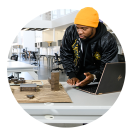 Student Nehemiah Ashford Carroll, wearing a black jacket and yellow beanie hat, stands in a studio in Lee Hall and gestures toward an architectural model project. 