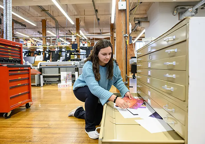 A female student wearing jeans and a light blue sweater kneels next to an open file cabinet drawer in a printing lab and looks at paper samples. 