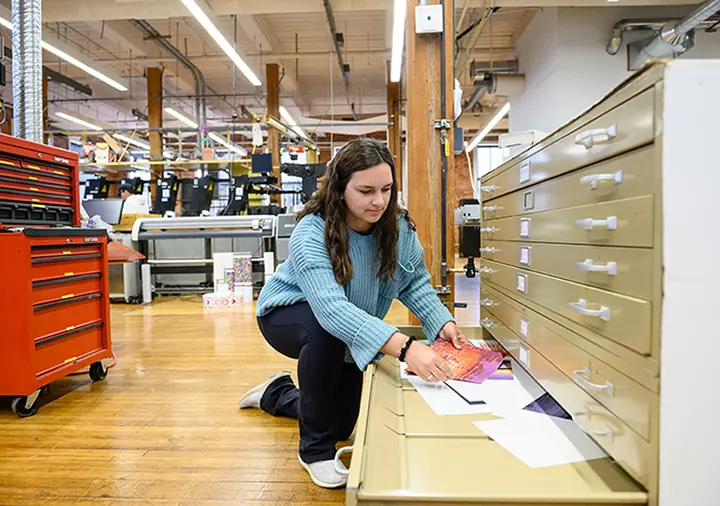 A female student wearing jeans and a light blue sweater kneels next to an open file cabinet drawer in a printing lab and looks at paper samples.