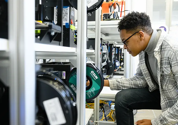 A male student wearing jeans, a gray turtleneck and a cream-and-gray plaid jacket leans next to some machinery and examines a roll of label tape.