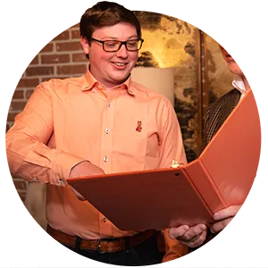 A male student wearing an orange-and-white checkered button-down and a male professor wearing a plaid tweed jacket look at the contents of an orange binder.