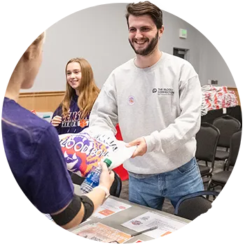 A male student stands behind a table with informational flyers about a blood drive while handing a sweatshirt that says “Blood Bowl” with a graphic of a tiger on it to a female student. 