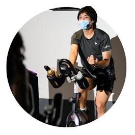 Student Hoang Nam Huynh, wearing black athletic attire and a blue face covering, uses an elliptical while leading a workout session. 
