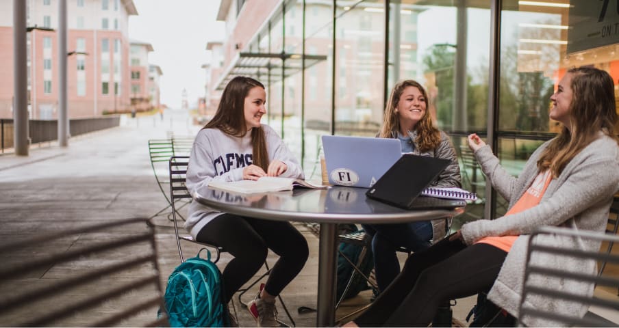 Three female students study and talk at a table outside the student bookstore in Douthit Hills.