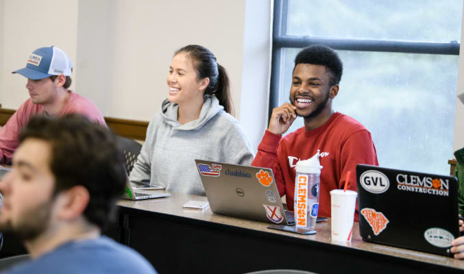Clemson business students smile during class in Sirrine Hall, sitting behind their stickered laptops.