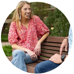 A female student in a pink and white blouse and light-wash blue jeans and a male professor wearing a light blue button-down shirt and blue jeans sit together on a brown wooden bench outside a brick academic building. 