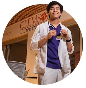 A male student wearing a white coat and a stethoscope stands in front of a sign for the Clemson School of Nursing.