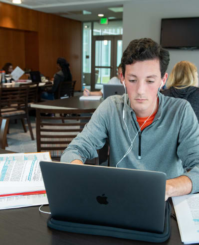 Male student wearing headphones works on his laptop with notes beside him on a table in Starbucks.