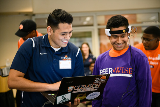 Industrial engineering student Patrick Mispireta smiles while helping a younger Hispanic male use wearable technology.