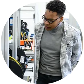 A male student wearing a plaid jacket, gray turtleneck and blue jeans observes laser-cutting and 3D-printing equipment inside the on-campus Makerspace. 