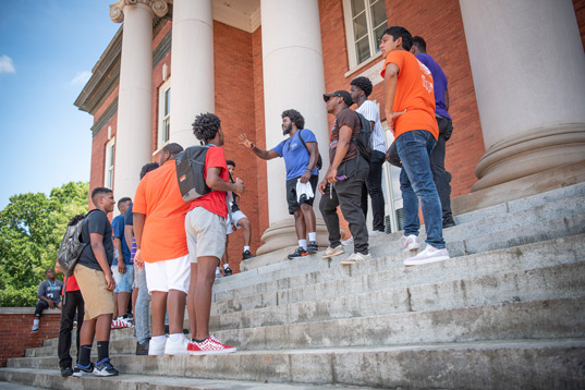 A group of diverse male high school students stands in a circle on the steps of Sikes Hall, listening to their campus guide.
