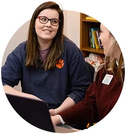 A female student wearing a blue pullover with an orange Tiger paw sits in front of a laptop and speaks with a female staff member. 